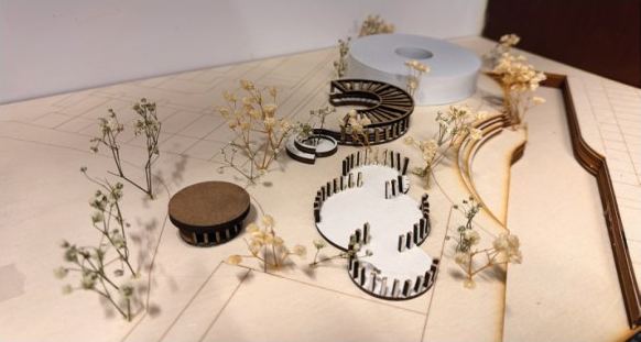 Photograph of the 1:200 site model from above: a white cylindrical hall, curved slatted elements and dried-flower trees on a plywood base.