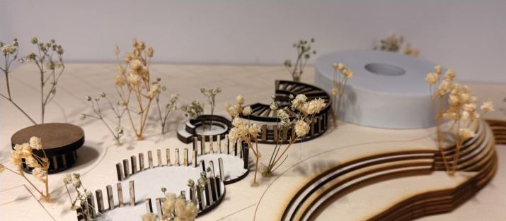 Wide view of the 1:200 site model: curved stacked plywood terraces sweep past slatted landscape elements and dried-flower trees to a white hall.