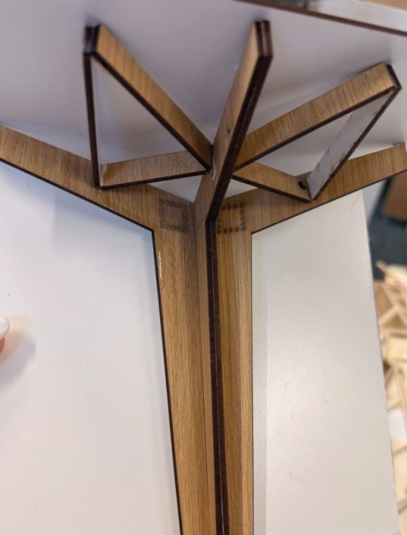 Photograph of a 1:20 tectonic model: a laser-cut timber column branching into angled arms with white card infills beneath a white roof board.