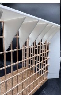 Close view of the tectonic model: triangular white card fins hanging from the roof edge over a square timber-stick lattice.